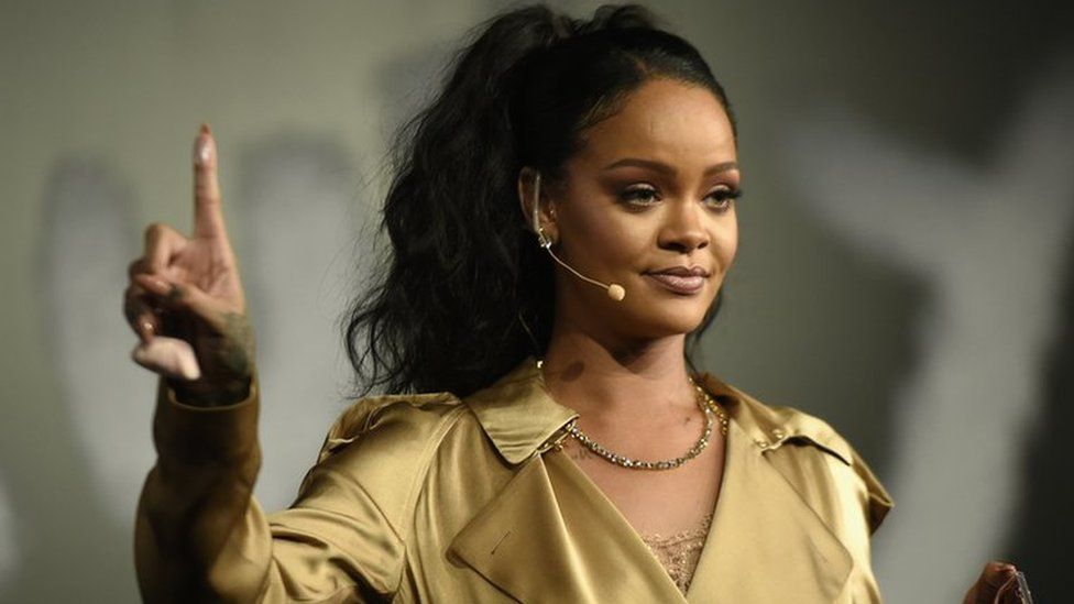 Rihanna wears a gold jacket and holds up one finger in a 'shush' motion